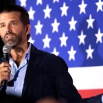 Man in a navy blazer holds a microphone and speaks on stage with a large American flag behind him behind him.