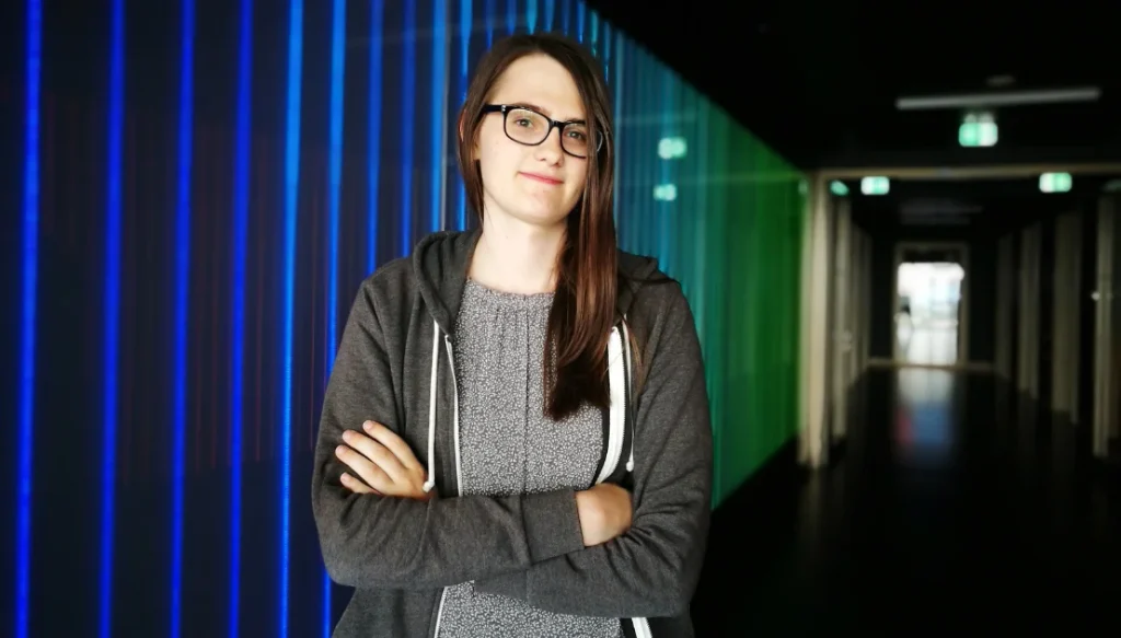 lilit-vittmann-2 » GP Woman with glasses and brown hair stands with arms crossed in a hallway lined with blue glass panels.