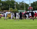 Youth football game on a sunny field; players in yellow and white uniforms gather near the sideline as spectators watch nearby.