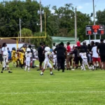 Youth football game on a sunny field; players in yellow and white uniforms gather near the sideline as spectators watch nearby.