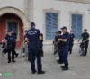 Police officers in navy uniforms with riot shields form a semicircle outside a building with blue shutters.