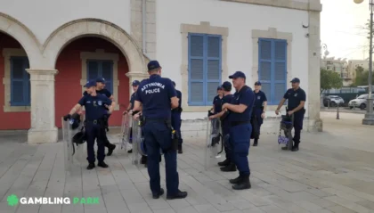 Police officers in navy uniforms with riot shields form a semicircle outside a building with blue shutters.