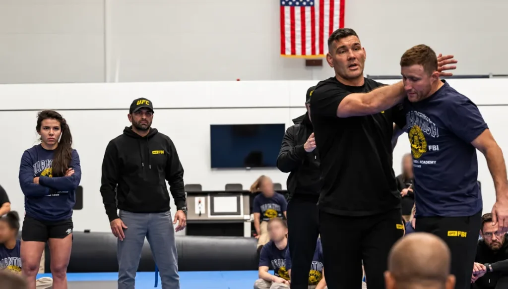 Instructor in a black shirt demonstrates a grappling hold on a male student while several people watch in a gym with an American flag on the wall.